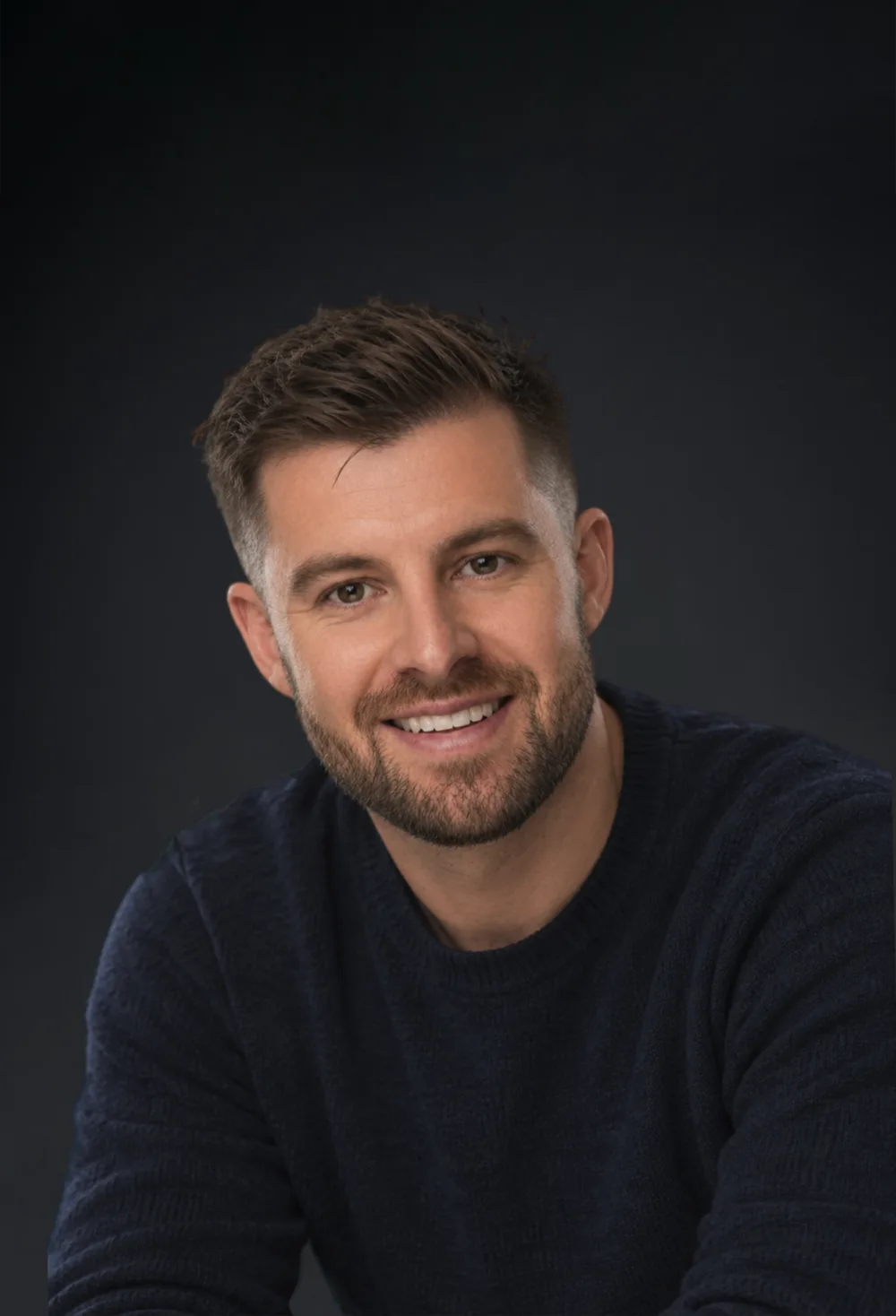 Fraser Macdonald, smiling, in a navy jumper against a dark studio background.