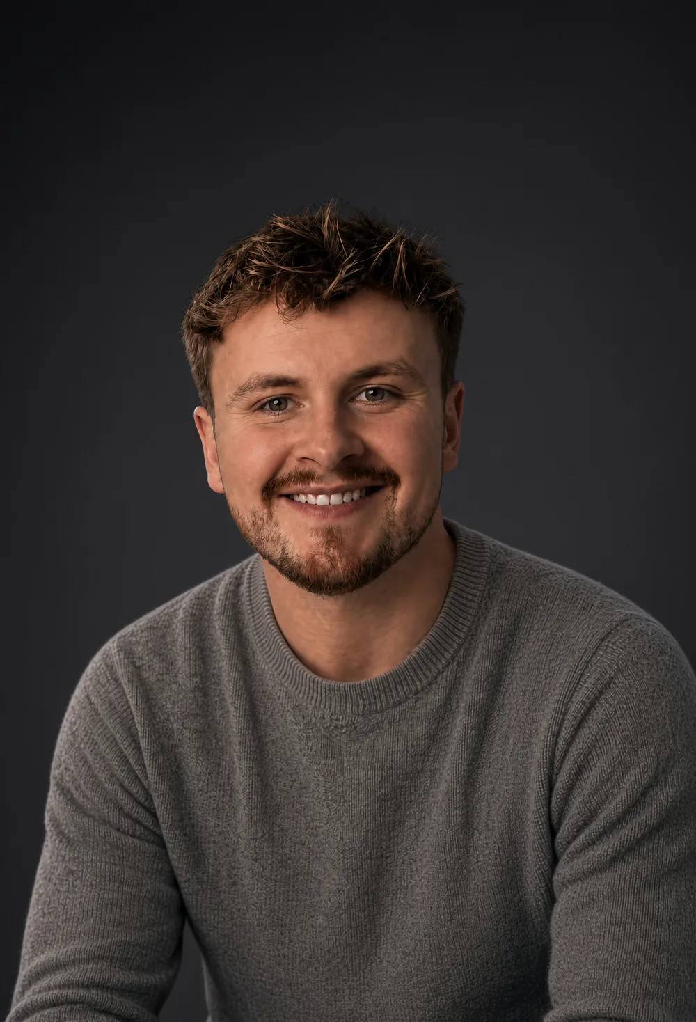 Oliver Cooper, smiling, in a grey jumper against a dark studio background.