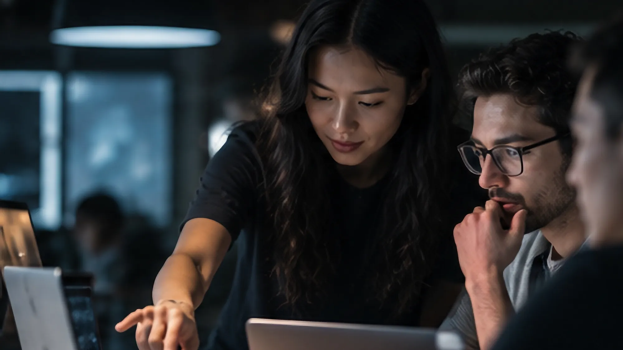 Three colleagues work together around laptops, with one pointing at a screen as the others observe.